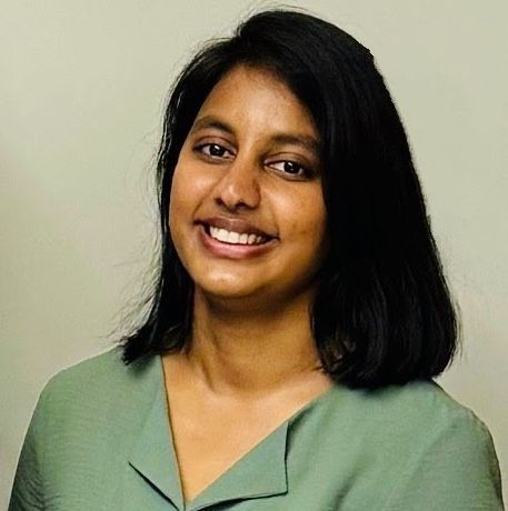 Person with shoulder-length dark hair wearing a light green top, standing against a neutral background.