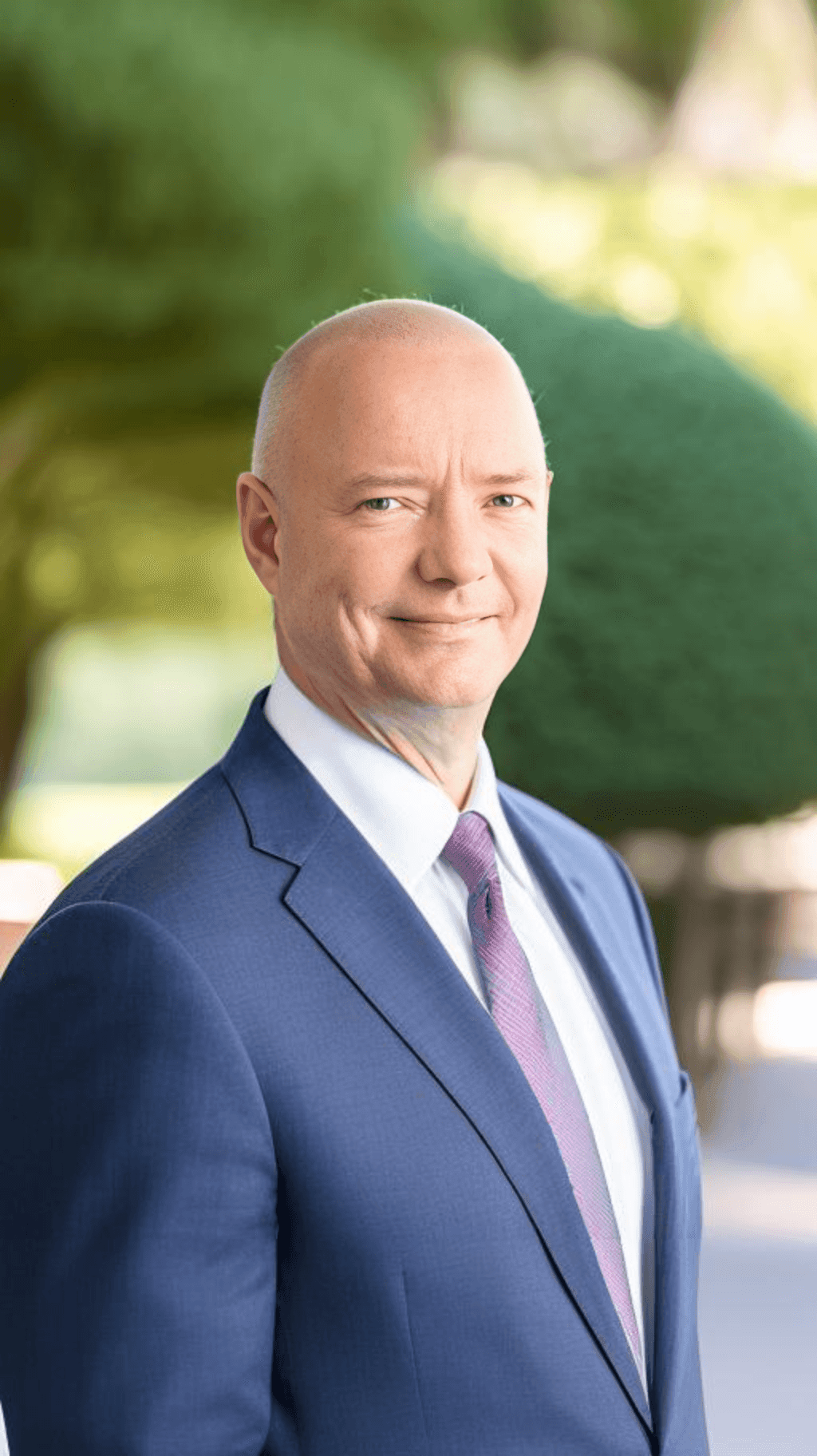 Man wearing a navy blue suit and pink tie, standing outdoors with blurred greenery in the background.