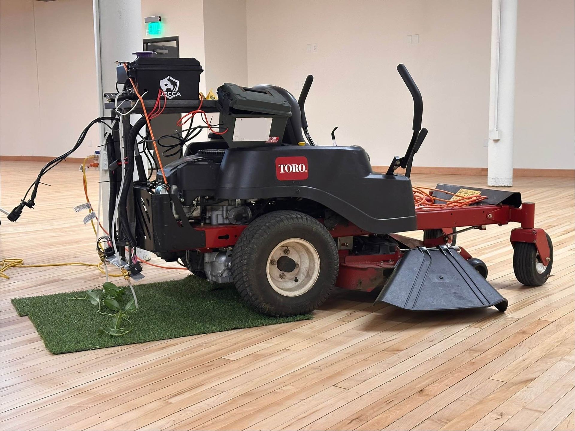 Modified Toro lawn mower with attached equipment inside a room, placed on a small grass patch.